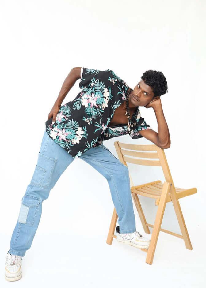 Casual men’s blue denim cargo pants with black floral shirt, model leaning on chair, showcasing side and knee pocket details and comfortable fit